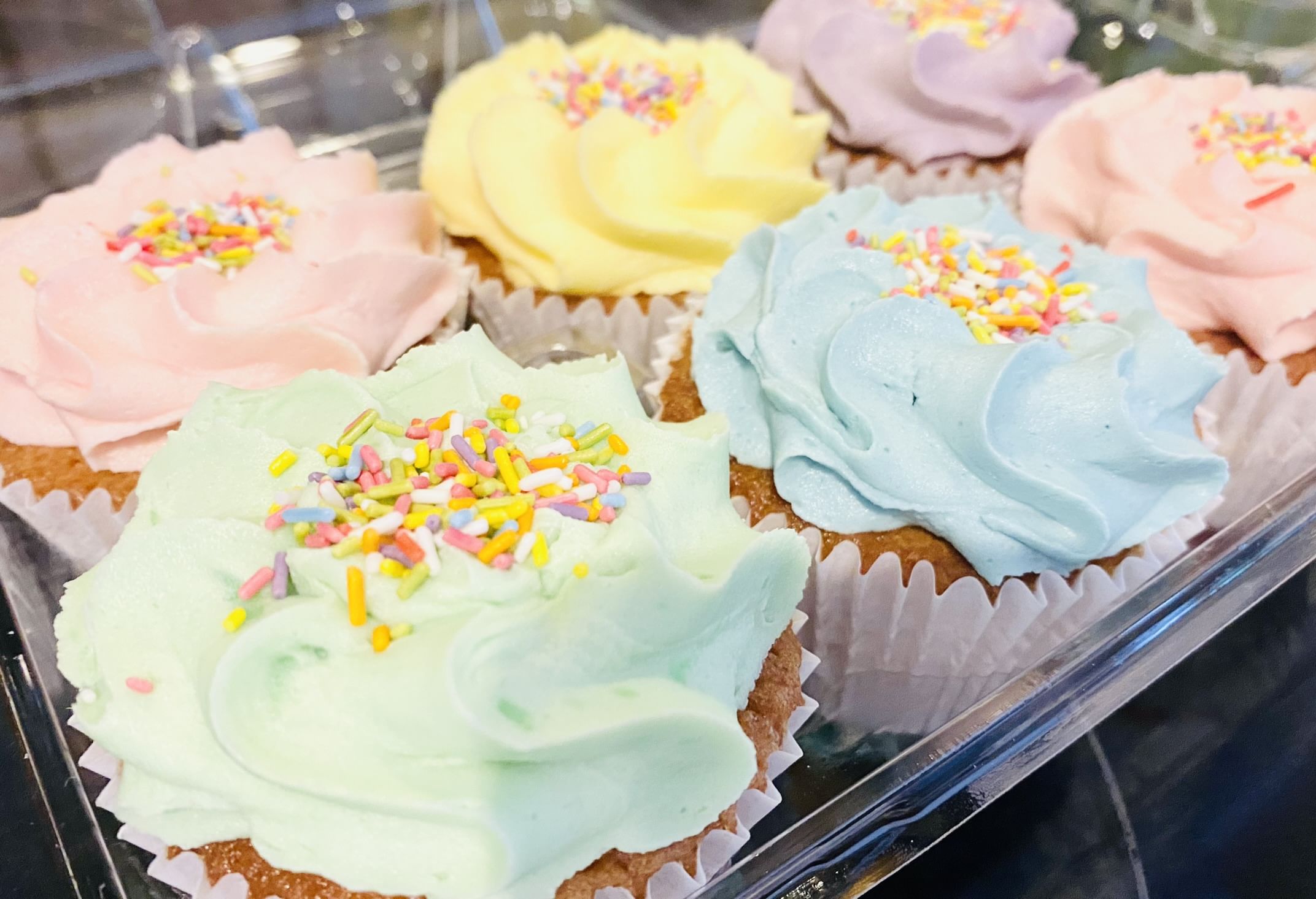 A close-up of six vanilla cupcakes displayed in a clear plastic container. Each cupcake is topped with colorful vanilla buttercream frosting in pastel shades of pink, yellow, purple, green, and blue, all sprinkled with multicolored sprinkles.